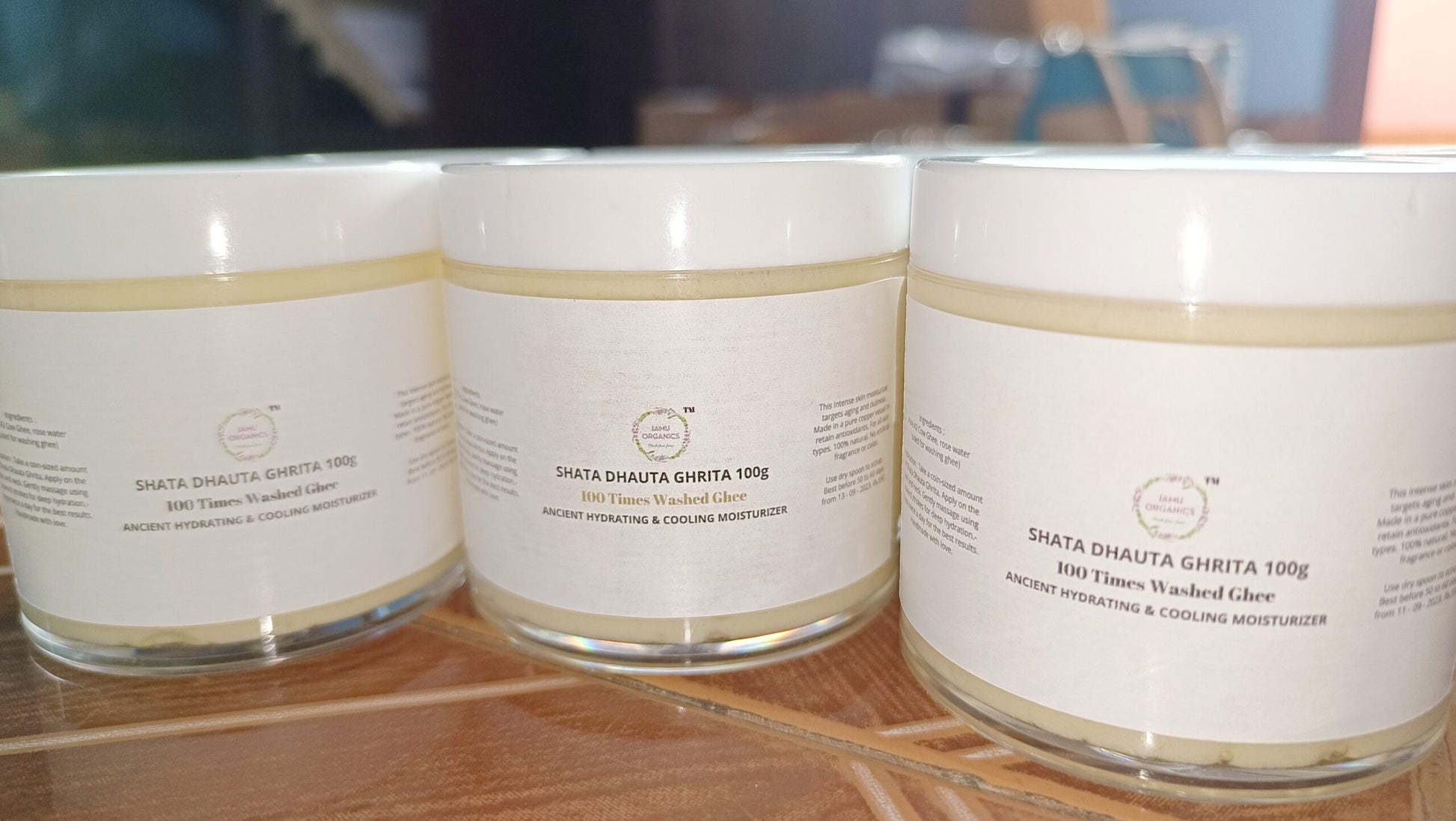 Three containers of Shata Dhauta Ghrita on a reflective surface with a blurred background.